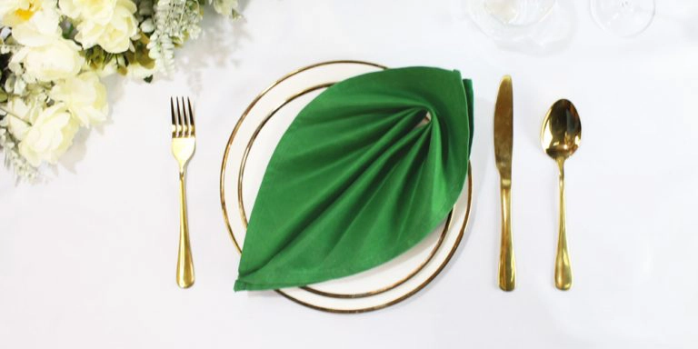 Napkin fold Leaf fold in a green napkin placed on a white plate with a gold rim